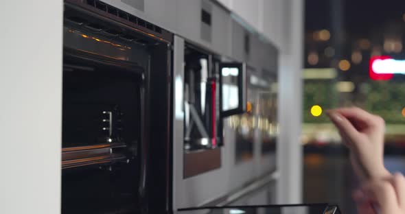 Woman Putting Baking Tray Into Electric Oven and Turning in on in Kitchen alt
