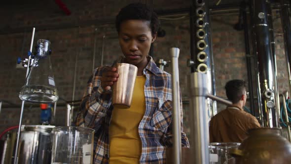 African american woman working at gin distillery smelling product and using tablet alt
