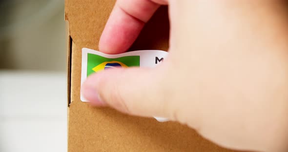 Hands applying MADE IN BRAZIL flag label on a shipping cardboard box with products. Close up shot wi alt