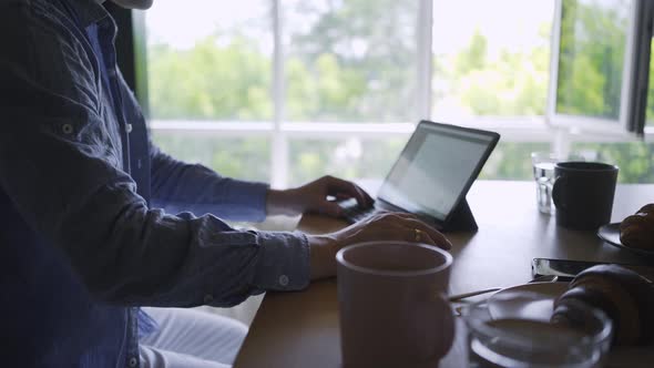 Man Works on Laptop at Table with Cup and Croissant in Room