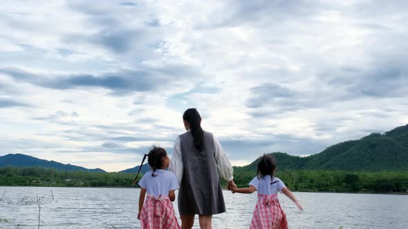 Happy mother walks hand in hand with cute little sisters enjoying the nature by the lake. alt