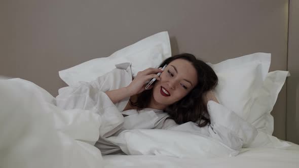 Young Joyful Woman Is Talking By Cellphone Lying in Bed at Morning at Weekend Chatting Cheerfully alt