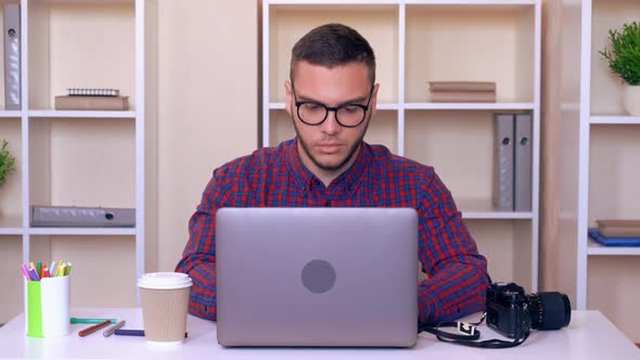 Man Wearing Bright Shirt Sitting at the Working Place Using Laptop alt