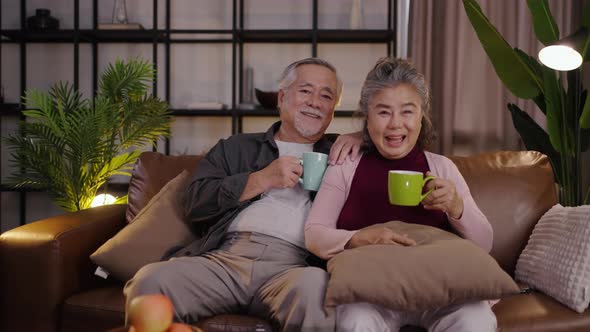 Elderly Asian couple watching a movie on their television at home at night while having a drink.