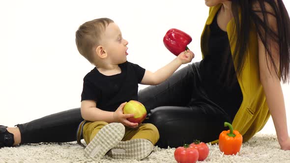 Mom and Son Are Sitting on the Floor Holding Vegetables. White Background alt