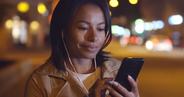 Bokeh Shot of African Woman in Earphones Using Smartphone Outdoors o in Night City alt