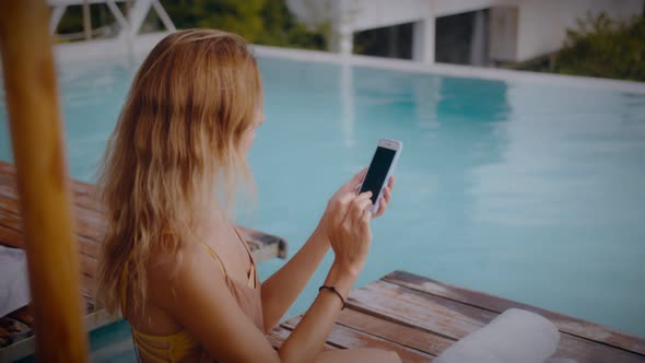 A Blonde Woman Uses Her Mobile Phone While Lounging in the Sun By the Pool on Vacation alt