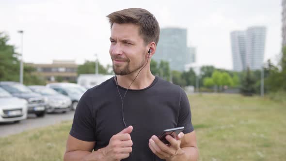A Young Man Listens To Music on a Smartphone with a Smile By a Parking Lot - City in the Background alt