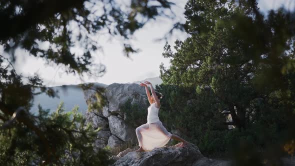Young Woman in Long White Dress Stands in Yoga Pose on Cliff