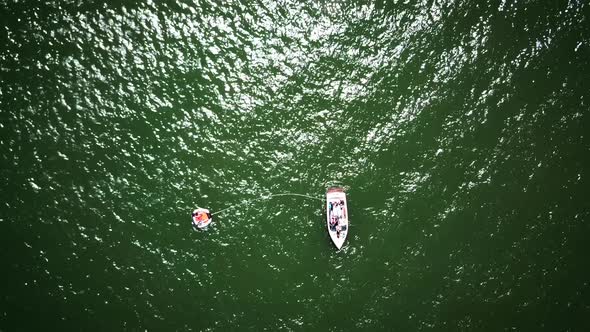 Drone shot zooming in on a boat staying still on a green lake. The boat has a rope leading to a tube alt