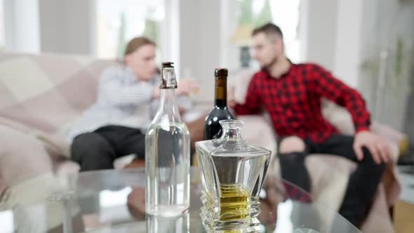 Full and Empty Alcohol Bottles on Glass Table with Two Blurred Men Clinking and Toasting at alt
