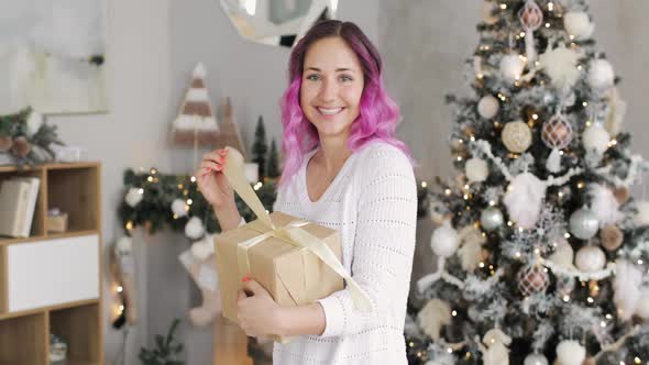 Young Happy Woman with Pink Hairs and Gift Box in Hands Near Christmas Tree