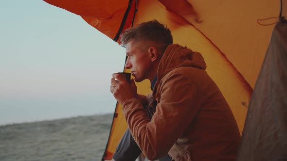 Adult Man Hiker Sitting at Tent Entry and Holding Mug with Coffee in Hands alt