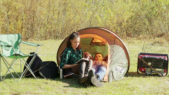Boyfriend Laying Down on Camping Tent While Girlfriend's Reading a Book alt