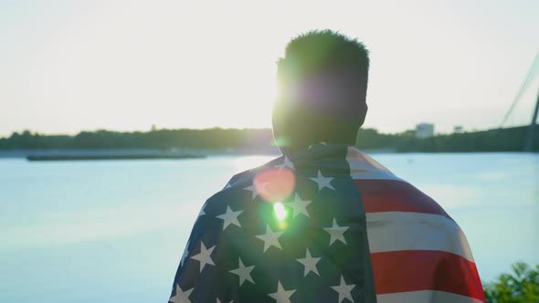 Afroamerican Man with American Flag on Shoulders Looks Into Distance at Sunrise alt