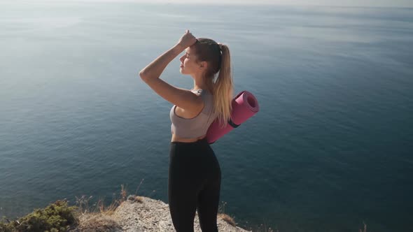 Woman Holds Yoga Mat, Looks at Sea From Cliff, Breathes Deeply, Slow Motion alt