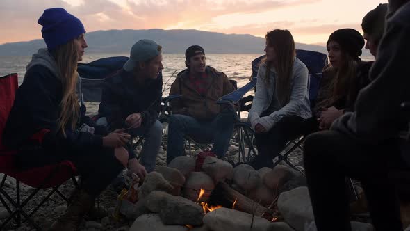 Group of family and friends gathered around campfire during sunset alt