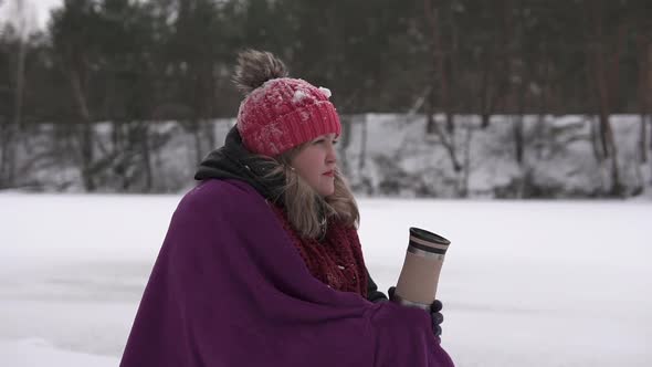 A beautiful girl in a winter forest near a frozen lake drinks tea from a thermo mug. alt