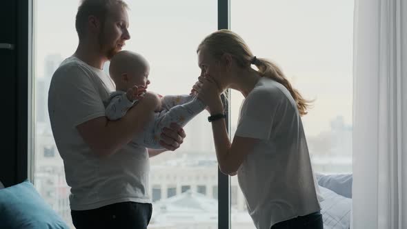 young parents with toddler play, kiss standing by the window