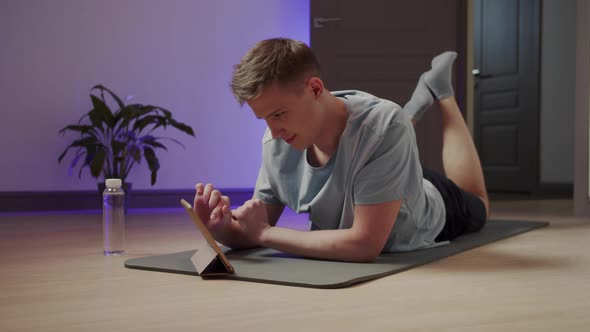 A Man Lies on a Yoga Mat and Watches Exercises on a Tablet alt
