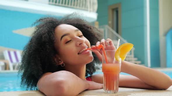 African American Woman in Red Swimsuit Drinking Non-alcoholic Orange Cocktail From Straw, Relaxation alt