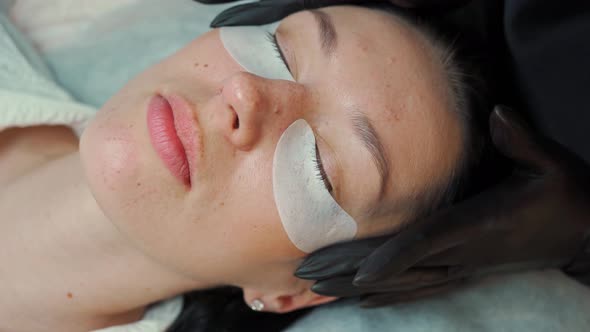 Eyelash Extension Master in Black Gloves Sticks Protective Patches Under the Client's Eyes Before alt