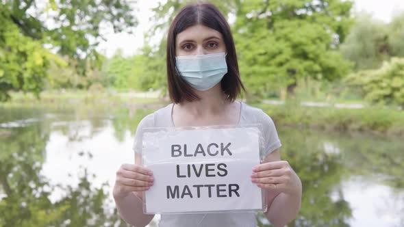 A Young Caucasian Woman in a Face Mask Shows a Black Lives Matter Sign to the Camera in a Park alt