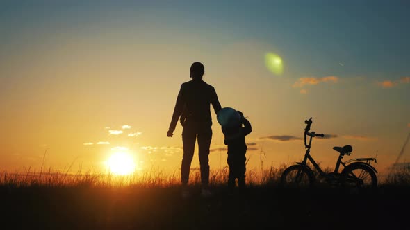 Silhouette of Mother Teaching Little Son to Ride a Bike at Meadow During Sunset alt