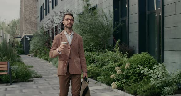 Cheerful Good Looking Guy in Glasses Carrying Bag and Coffee Paper Cup. Stylish Bearded Businessman alt