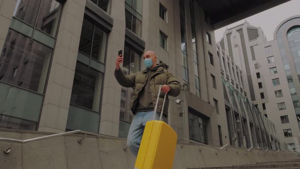 European Man Coming Out of Office Center with Yellow Suitcase and Talks Facetime in Protective Mask alt