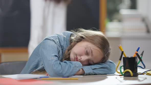 Tired Caucasian Schoolgirl Sleeping on Desk in Classroom with Blurred Classmate Jumping at alt