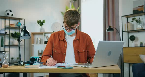 Young Male Freelancer or Worker in Facial Medical Mask Working on Laptop at Home, Covid-19 alt