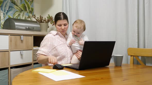 Woman holding a baby and working on her laptop, taking notes on paper