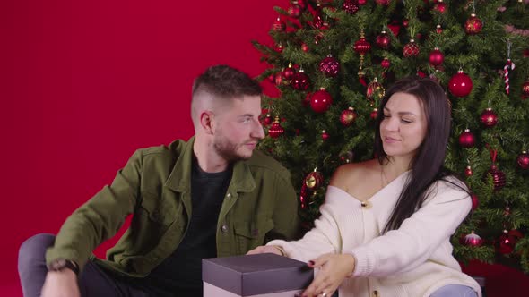 Young Loving Couple Opening Long Awaited New Year Gift Sitting Near Xmas Tree