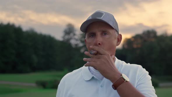 Senior Golf Player Smoking Cigar Looking Camera on Sunset Field Course Fairway alt