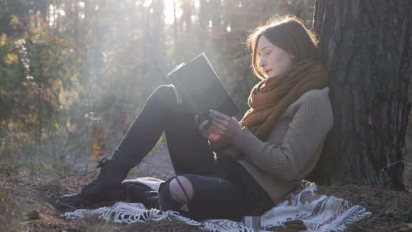 Happy charming woman traveler sitting on plaid, reading book and relaxing in autumn forest at sunset alt