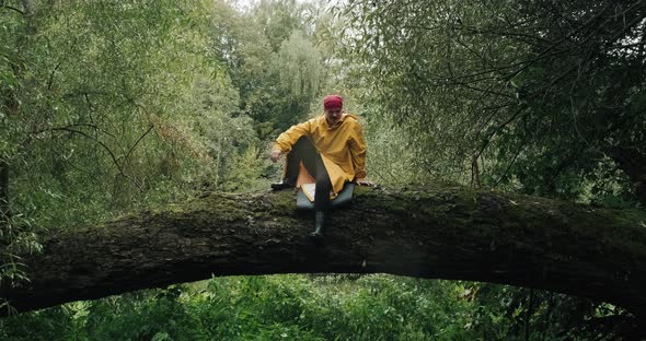 Tourist in a Yellow Raincoat Drops a Backpack From a Tall Trunk of a Fallen Tree