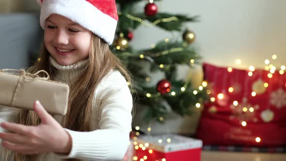 Someone Gives Little Girl Gift Box on the Background of Christmas Tree alt