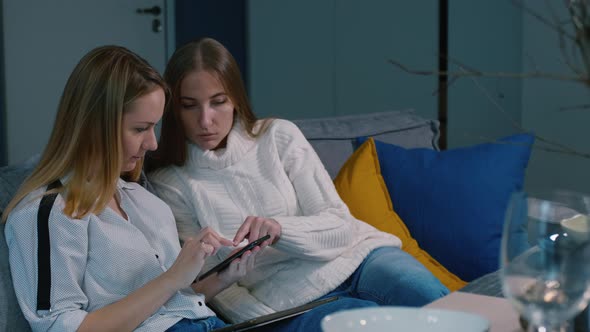 Young Cheerful Women Sitting on Sofa at Dinner Table and Using smartphone