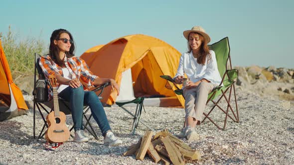 Couple Charming Beautiful Women Girlfriends Sitting in Camping Chairs at Sea Coast Have Fun Talking alt