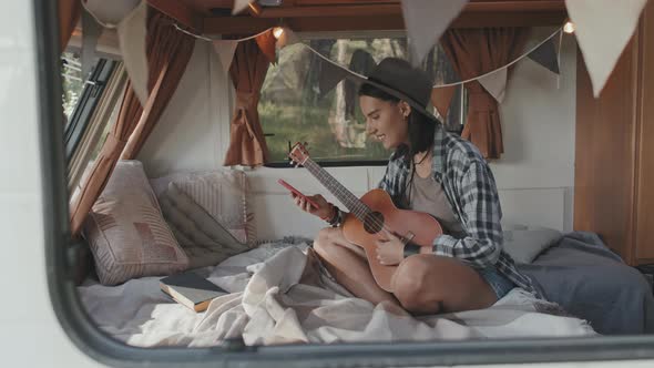 Happy Woman with Ukulele Using Mobile Phone in Camper alt