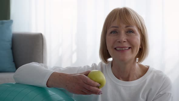 Senior Lady Holding Apple Sitting Near Fitball After Workout Indoors alt