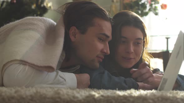 Young Guy and Girl Covered with a Blanket Lying on the Floor Looking at Laptop at Home alt