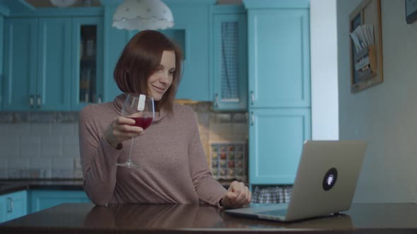 Young 30s smiling woman drinking glass of red wine and typing on laptop alt