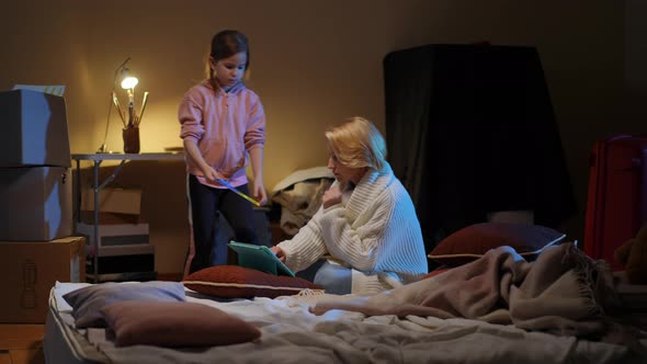 Wide Shot Ukrainian Girl Showing Placard to Woman Hiding with Parent in Bomb Shelter alt
