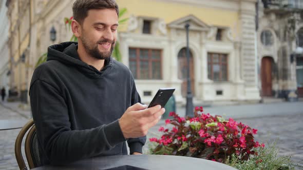Hipster Guy with Mobile Smart Phone Digital Camera Talks While Sitting in Cafe alt