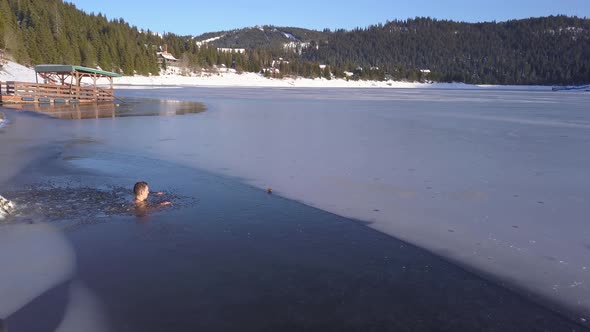 Aerial views of man sitting neck deep in frozen mountain lake, drone rotating. Young guy doing polar alt