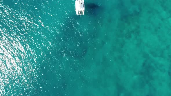 A bird's-eye view of a lone white catamaran sailboat sailing along on the tranquil clear turquoise o alt
