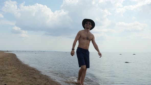 Shirtless Man in Cowboy Hat and Sunglasses Walking on Sandy Beach