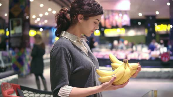 Young Beautiful Brunette Girl in Her 20's Picking Out Bananas and Putting Them Into Shopping Cart at alt